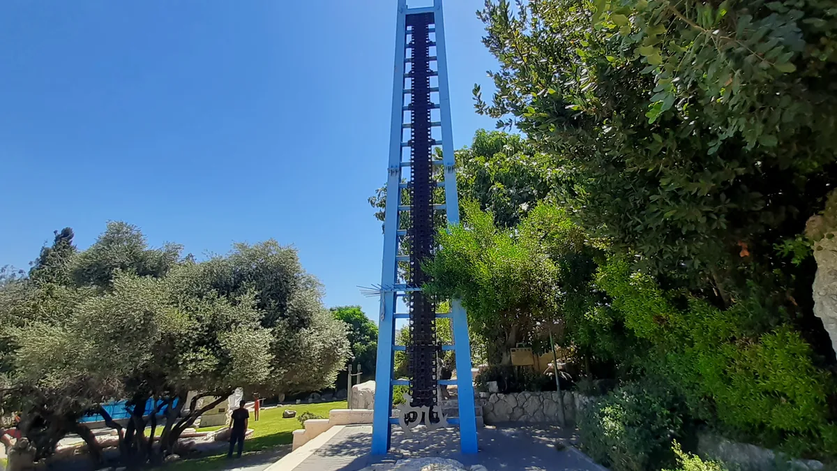 A tall blue steel sculpture among trees and lawn in the Gan Golan sculpture garden