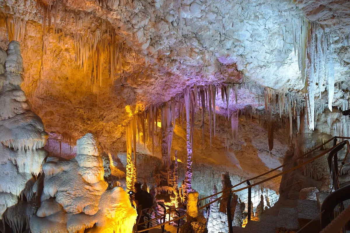 Lit stalactites and stalagmites inside the Stalactite (Avshalom / Soreq) Cave, with the visitors' walkway and railings on the right