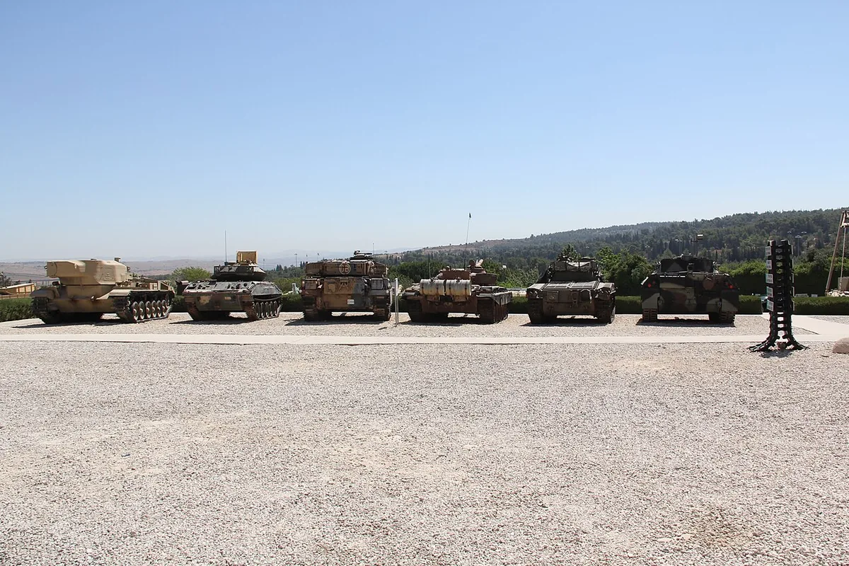 A row of tanks on display on the gravel parade ground of the Yad La-Shiryon armoured corps museum at Latrun, with wooded hills behind
